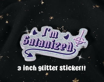 May include: A 3-inch glitter sticker with the text "I'm Satanized" in purple and silver. The sticker has a unique shape with a stylized symbol and crescent moon accents. The background is black with gold glitter.