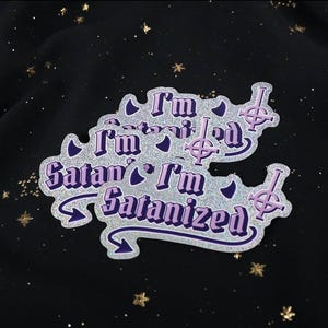 May include: A set of glittery stickers with the text "I'm Satanized" in purple and white. The stickers feature a stylized cross symbol and are set against a dark background with star-shaped sparkles.