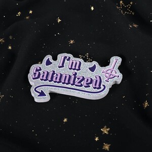 May include: A glittery, die-cut pin with the text "I'm Satanized" in purple and white. The pin features a stylized arrow and a cross symbol, set against a black background with gold star accents.
