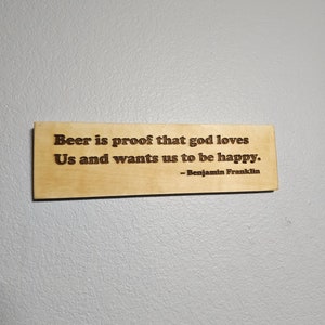 May include: A wooden sign with a quote about beer and happiness. The quote reads "Beer is proof that god loves Us and wants us to be happy." The quote is attributed to Benjamin Franklin.