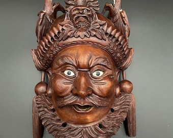 May include: A detailed, hand-carved wooden mask with a stern expression. The mask features a prominent moustache, open mouth, and white eyes. Intricate carvings adorn the forehead and sides, with decorative elements including dragons and a crown. The mask is a rich brown colour.