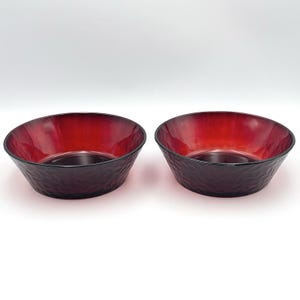 Puede incluir: Dos cuencos redondos a juego hechos de vidrio rojo intenso. Cada cuenco presenta una superficie exterior texturizada y martillada con un acabado interior liso y brillante. El vidrio pasa de un carmesí oscuro saturado a un borde rojo más claro.