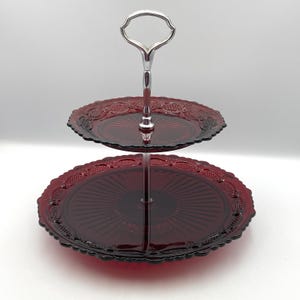 Op de afbeelding: Een serveerschaal met twee niveaus en een dieprood glazen ontwerp. De borden hebben een geschulpte rand en een radiaal patroon. Een zilveren metalen handvat verbindt de twee niveaus.