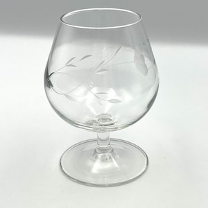 May include: Clear brandy snifter with etched floral design. The glass features a rounded bowl, a short stem, and a wide base. Delicate etched leaves and a flower adorn the glass, adding a touch of elegance. Perfect for serving brandy or other spirits.