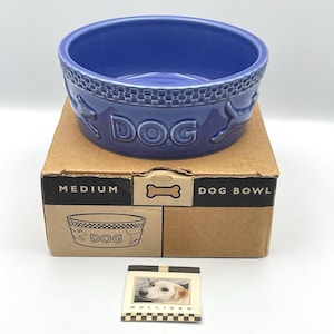 Peut inclure: Un bol pour chien en céramique bleu moyen avec un bord à carreaux, des silhouettes de chien en relief et le mot DOG. Il repose sur une boîte en carton marron marquée MEDIUM DOG BOWL avec une icône d'os. Une carte montre un chien et MULLIGAN.