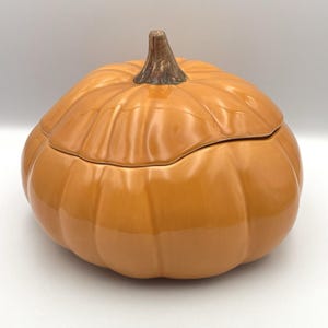 May include: A ceramic pumpkin-shaped container with a lid. The container is a glossy orange color with a brown stem. The lid fits snugly on top, creating a complete pumpkin shape. This decorative piece is perfect for fall and Halloween.