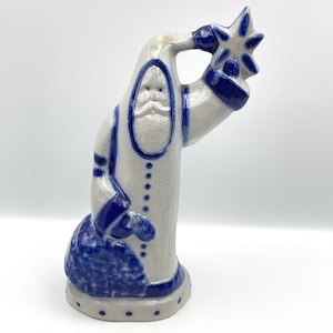 May include: A ceramic Santa Claus figurine in a whimsical design. The figure is primarily off-white with blue accents, including the trim, cuffs, and a star held in one hand. The face has a detailed beard and a smiling expression. The figurine is approximately 8 inches tall.