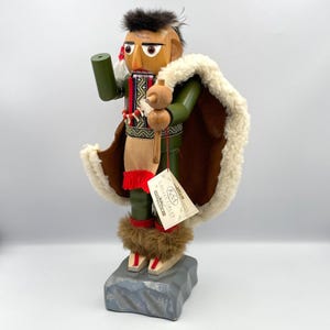 Puede incluir: Figura de cascanueces de madera con diseño nativo americano. La figura lleva una chaqueta verde, una capa marrón con ribete blanco y botas de piel sintética. Sostiene una herramienta de madera y un cilindro verde. Una etiqueta cuelga de la figura.