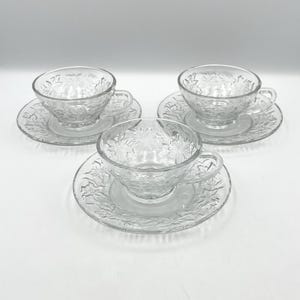 May include: Three clear glass teacups and matching saucers, each featuring a delicate embossed floral pattern across the surface. The cups have a single curved handle and a smooth interior, while the saucers have a matching textured rim.