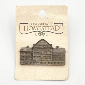 Könnte beinhalten: Ein Longaberger Homestead-Anstecknadel mit einem bronzefarbenen Crawford Barn-Design. Die Anstecknadel ist an einer cremefarbenen Karte mit dem Longaberger Homestead-Logo befestigt. Die Anstecknadel ist eine detaillierte Miniatur einer Scheune.