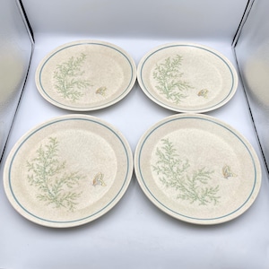 May include: A set of four round ceramic plates with a speckled beige base. Each plate features a light blue rim, a green fern-like botanical illustration on the left side, and a small yellow and blue butterfly graphic on the right side.