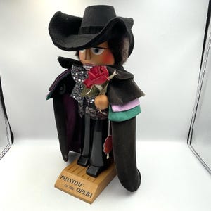 May include: A Phantom of the Opera nutcracker figurine. The figure wears a black hat, cape, and suit, and holds a red rose. The base is wooden and has the words "Phantom of the Opera" engraved on it.