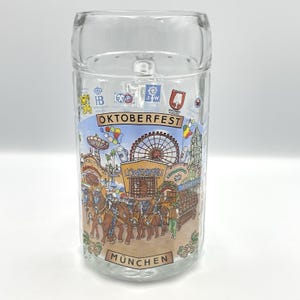 May include: Clear glass beer stein with a handle. The stein features a colourful illustration of the Oktoberfest celebration, including a Ferris wheel, horses, and the text "OKTOBERFEST" and "MUNCHEN".