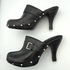 May include: Pair of black leather clogs with high heels. Each clog features a strap with a silver buckle, silver stud accents, and a wooden heel. The shoes are designed for a stylish, elevated look.