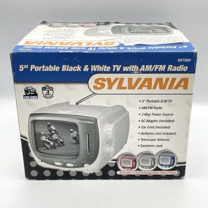 Könnte beinhalten: Ein tragbarer 5-Zoll Schwarz-Weiß-Fernseher von Sylvania mit AM/FM-Radio, verpackt in einem blau-weißen Karton. Die Verpackung zeigt das Produkt, Funktionen wie eine 3-Wege-Stromversorgung und drei austauschbare Frontblenden in Rot, Grau und Blau.