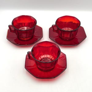 Può includere: Tre tazze da tè con piattini abbinati in vetro rosso rubino. Ogni tazza presenta un design ottagonale sfaccettato con un manico robusto, mentre i piattini corrispondenti riprendono la stessa forma a otto lati.