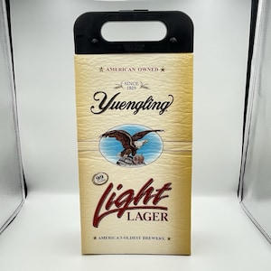 Pode incluir: Um porta-cervejas de cartão com uma alça preta. A frente apresenta o logotipo Yuengling, um gráfico de águia e as palavras "light lager". O texto também inclui "American Owned" e "America's Oldest Brewery."
