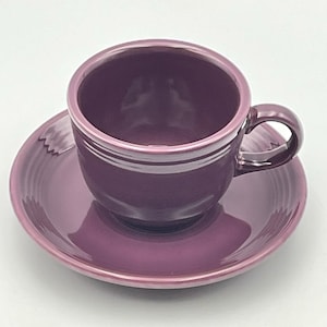 Könnte beinhalten: Ein tiefviolettes Tassen- und Untertassen-Set aus Keramik mit glänzender Oberfläche. Die Tasse hat einen abgerundeten Körper mit einem einzelnen geschwungenen Henkel an der Seite, während die Untertasse konzentrische Rillen am Rand aufweist.