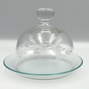 May include: A clear glass cloche with a matching plate. The dome-shaped cover features etched floral designs and a rounded handle. The plate has a slightly raised edge. Ideal for serving cheese or desserts.