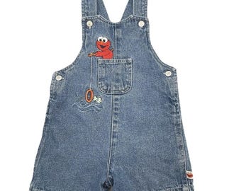 May include: Blue denim overall shorts featuring an embroidered Elmo character fishing. The shorts have adjustable straps, a front pocket, and button closures. The Elmo design includes a fishing rod, a fish, and waves.