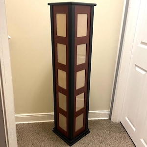 May include: A tall, square, floor-standing photo display tower featuring a dark red frame with black trim. The structure includes five vertical rectangular openings on each visible side, designed to hold photographs behind clear panels.