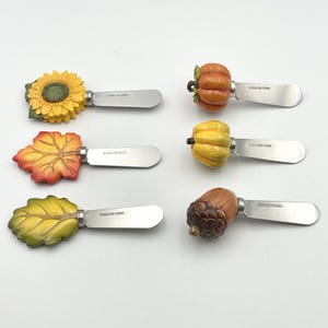 May include: Set of six decorative cheese spreaders with stainless steel blades. The handles feature autumn-themed designs, including a sunflower, leaf, pumpkin, and acorn. The blades are approximately 4 inches long, perfect for serving cheese and other spreads.