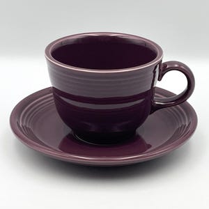 Könnte beinhalten: Ein tiefviolettes Keramik-Teeservice bestehend aus einer Tasse und einer passenden Untertasse. Die Tasse hat einen abgerundeten Körper mit horizontalen Rillen an der Außenseite und einen geschwungenen Henkel. Die Untertasse hat einen gerillten Rand.