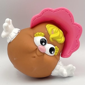 Pode incluir: Um brinquedo castanho, em forma oval, com uma cobertura de cabeça rosa e cabelo amarelo. O brinquedo tem olhos grandes e expressivos, um nariz rosa e mãos brancas. O brinquedo é um personagem caprichoso, provavelmente um brinquedo infantil.