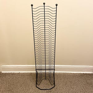 以下が含まれることがあります： 波型のデザインが施された、背の高い黒い金属製のマガジンラック。ラックは長方形のベースで、3つの装飾的なボールフィニアルが付いています。雑誌やその他の読み物を収納するように設計されています。