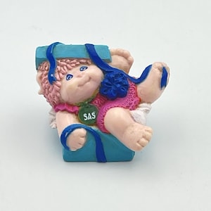 Peut inclure: Figurine Cabbage Patch Kids dans une boîte bleue avec un ruban bleu. La poupée a des cheveux roses, des yeux bleus et porte une tenue rose. Une étiquette verte avec "SAS" est attachée à la poupée.