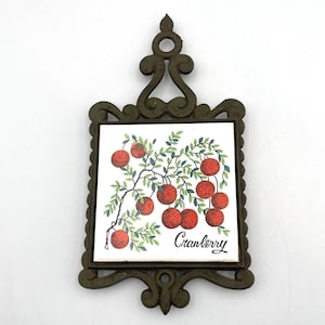 Puede incluir: Salvamanteles decorativo de metal con una baldosa de cerámica cuadrada con una ilustración de arándanos. La baldosa muestra arándanos rojos en una rama con hojas verdes, y la palabra "Cranberry" en escritura cursiva. El marco de metal es verde oscuro.