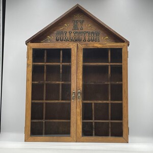 May include: A wooden wall-mounted display cabinet with a gabled roof shape, featuring two glass-paned doors with metal pulls. The top panel is inscribed with the text MY COLLECTION in black lettering with gold decorative flourishes.