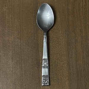 May include: A silver spoon with an oval bowl and a decorative handle. The handle features ornate, symmetrical designs and is rectangular in shape. The spoon is made of a shiny, metallic material and rests on a wooden surface.