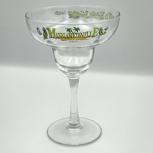 May include: Clear margarita glass with a wide bowl and a long stem. The glass features the text "Margaritaville Orlando" in green and yellow, along with tropical imagery. The glass is designed for serving margaritas.