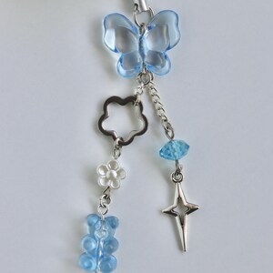 May include: A light blue butterfly charm with a silver chain, flower, and gummy bear accents. The charm is attached to a white earbud case. Additional charms include a star and a blue gem.