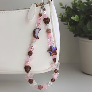 May include: A beaded bag charm with a variety of pink, brown, and clear beads. The charm includes heart, star, moon, and butterfly shapes. The charm is attached to a white purse with a gold-colored clasp.