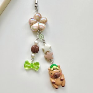 May include: A brown and tan capybara charm with a green turtle on its head. The charm is attached to a string of beads and a flower charm. The beads are white, brown, and green.
