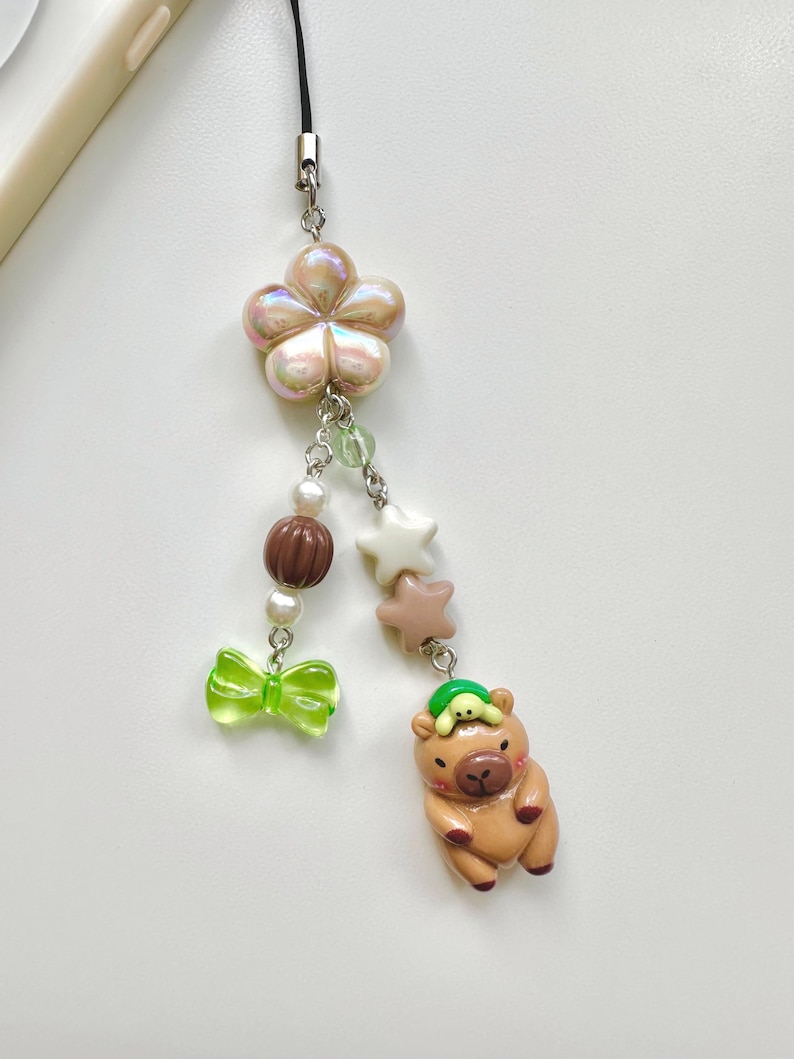 May include: A brown and tan capybara charm with a green turtle on its head. The charm is attached to a string of beads and a flower charm. The beads are white, brown, and green.