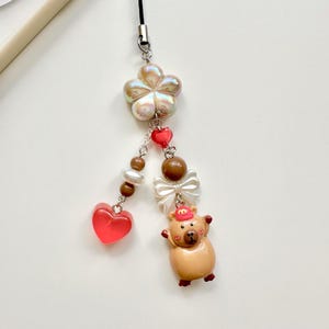May include: A brown and tan pig charm with a red hat and a white bow. The charm is attached to a black string with a white pearl flower, brown beads, and red hearts.