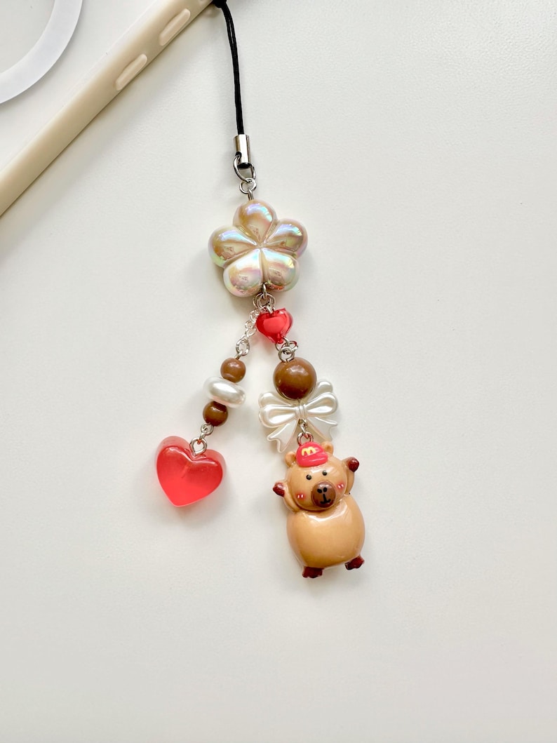 May include: A brown and tan pig charm with a red hat and a white bow. The charm is attached to a black string with a white pearl flower, brown beads, and red hearts.