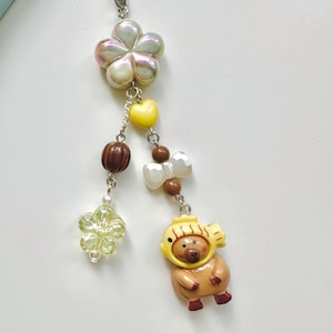 May include: A colorful phone charm with a brown bear wearing a yellow fish hat, a white pearl flower, a brown and white bow, a yellow heart, and a iridescent flower.