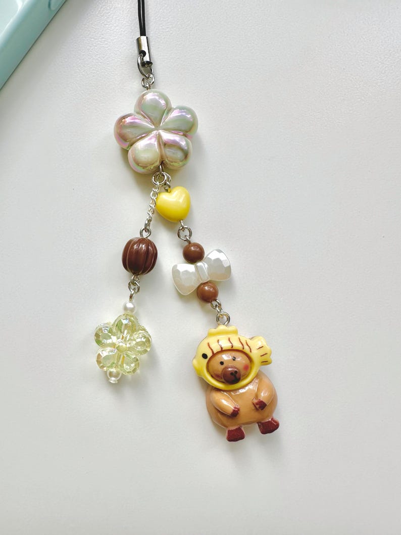 May include: A colorful phone charm with a brown bear wearing a yellow fish hat, a white pearl flower, a brown and white bow, a yellow heart, and a iridescent flower.