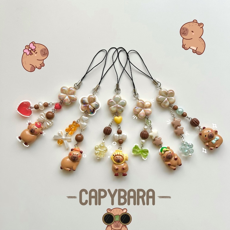 May include: Six colorful capybara charms with different accessories, including a heart, a bow, a flower, a star, and a hat. The charms are attached to black strings with beads and are hanging from a white background. The text "CAPYBARA" is written in black below the charms.