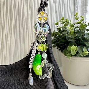 May include: A bag charm featuring a silver-tone lobster clasp, a yellow citrus slice charm, an iridescent bow, a green pickle charm, a star-shaped charm, and white letter beads spelling PICKLES, all suspended from a silver-tone metal chain.