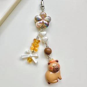 May include: A brown and tan capybara charm with a small orange pumpkin on its head. The charm is attached to a silver chain with a white flower, white hearts, and orange bear beads.
