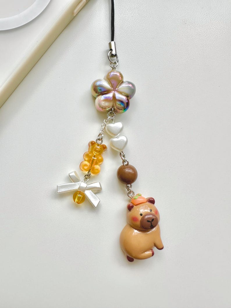 May include: A brown and tan capybara charm with a small orange pumpkin on its head. The charm is attached to a silver chain with a white flower, white hearts, and orange bear beads.