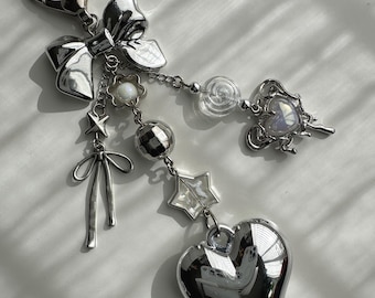 May include: A silver-toned keychain featuring a heart-shaped lobster clasp, a large metallic bow, and multiple dangling charms including a small flower, a disco ball bead, a star, a thin bow, a clear swirl bead, and a large reflective heart.