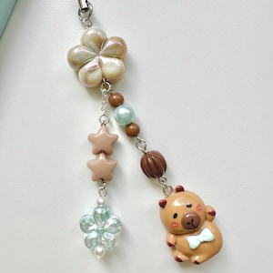 May include: A brown bear charm with a blue bow tie, a light blue flower charm, two pink star charms, and a pearl and brown bead charm on a black phone strap.