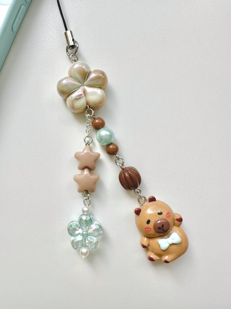May include: A brown bear charm with a blue bow tie, a light blue flower charm, two pink star charms, and a pearl and brown bead charm on a black phone strap.
