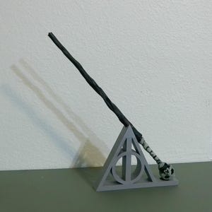 May include: A dark gray wand with a textured handle rests on a gray Deathly Hallows symbol stand. The wand has a detailed design, and the stand is triangular. The wand's tip has a white and black pattern.
